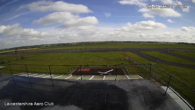 A wide view of a grassy airfield under a partly cloudy sky. In the foreground, there is a flat rooftop with dark floor. A railing runs along the edge of the roof. On the ground below, there is an area marked with a red square and a white "X". The airfield extends into the distance with patches of grass and a runway visible, along with a few scattered trees and a fence lining the sides. The overall scene conveys a calm and open atmosphere.