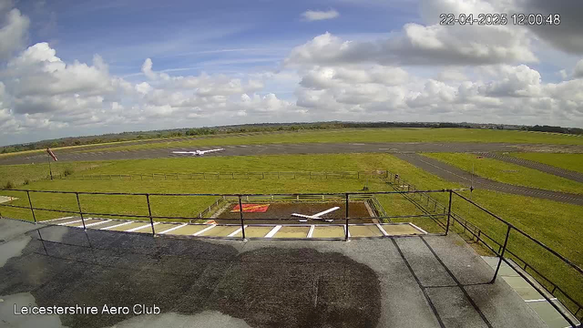 A view from a webcam at the Leicestershire Aero Club, showing a wide grassy field with an airplane on the runway. The sky is partly cloudy with blue patches, and there are some trees in the distance. A red windsock is visible on the left side of the image. The foreground shows a ledge or railing, and there is a white shape that appears to be the outline of an airplane on the ground.