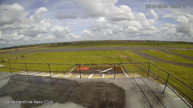 A view from a webcam at the Leicestershire Aero Club showing a grassy airfield under a partly cloudy sky. In the foreground, there is a railing with a puddle on the ground. The airfield is mostly clear with a small red square and a white cross marking a helipad area. In the background, the airstrip is visible with white lines for runway markings, surrounded by green fields and trees under a blue sky with scattered clouds. The time displayed is 13:00:13 on April 22, 2025.