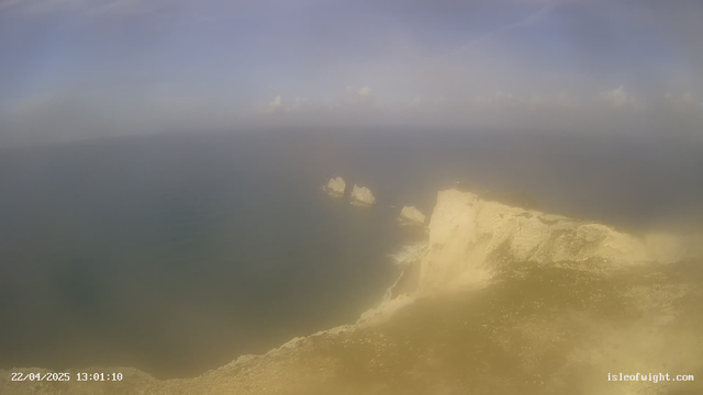 A hazy coastal view featuring rugged cliffs on the right side of the image, with the sea stretching out to the horizon. There are several rocky outcrops visible in the water. The sky appears mostly clear with some clouds. The date and time are displayed in the lower left corner of the image.