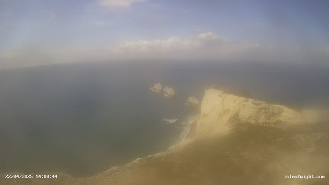 A panoramic view of a coastal area featuring cliffs and the sea. The cliffs are light-colored and rugged, dropping steeply into the blue water below. In the distance, there are several rocky outcrops emerging from the water. The sky is mostly clear with a few scattered clouds. The image appears to have a slight haze, suggesting a sunny day. The date and time are displayed in the lower left corner.
