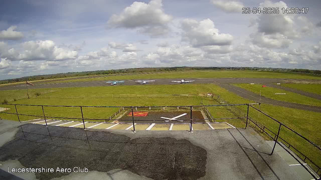 A view from a webcam at Leicestershire Aero Club shows a wide, grassy airfield under a partly cloudy sky. In the foreground, there is a paved area with an emblematic white cross and a small rectangular patch. Three small aircraft are parked on the runway, and a visible red flag indicates an area of caution. The background features rolling green hills and a clear horizon. The overall atmosphere is calm and sunny.