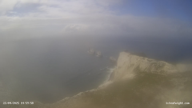 A coastal scene featuring a cliff edge with a rocky shoreline. The water is visible, along with several large rock formations in the sea. The sky is partly cloudy, with some blue peeking through. The image appears to have a hazy or foggy quality, obscuring details in the distance. A date and time stamp is located at the bottom left corner of the image, indicating it was taken on April 22, 2025, at 14:59:58.