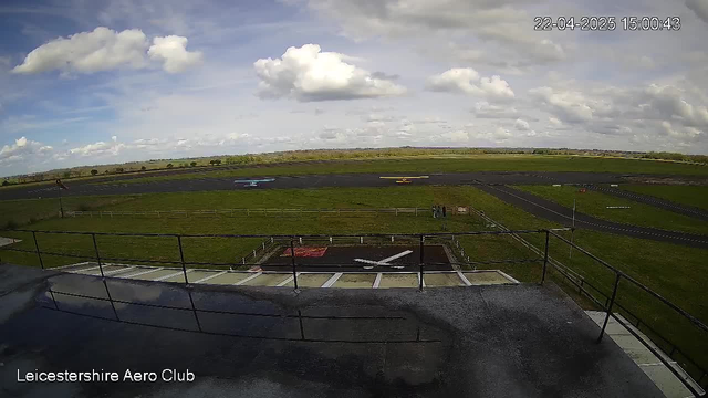 A view from a webcam at Leicestershire Aero Club shows a grass runway with several small planes, one blue and another yellow, parked on the tarmac. There is a grassy area with a few people standing near a fenced section. The sky is partly cloudy with scattered clouds and a serene atmosphere. The image is captured during the daytime.