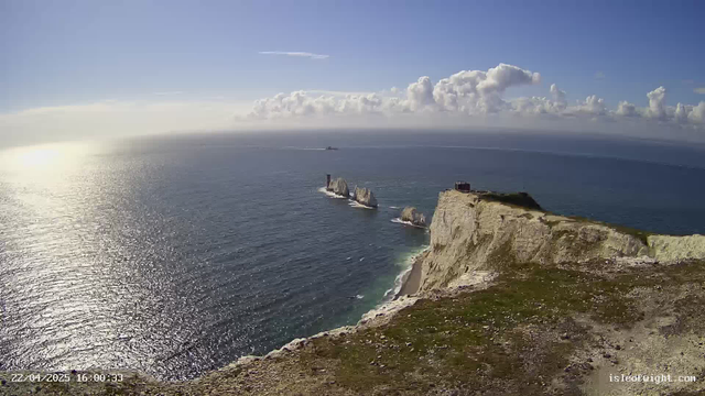 A panoramic view of the sea and coastline under a clear blue sky. The sunlight reflects off the water, creating a shimmering effect. In the foreground, a rugged cliff is visible, covered with grass and rocks. Several large white rock formations emerge from the water, and in the distance, a small boat is visible on the horizon. Fluffy white clouds decorate the sky, adding depth to the serene landscape.