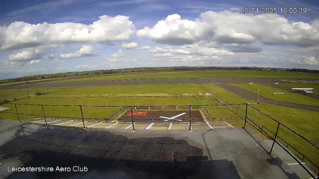 A view from a webcam at Leicestershire Aero Club showing a wide, open landscape. The foreground includes a helipad marked with a white "X" on a gravel surface. Behind the helipad, there is a grassy area and a runway, which is mostly paved. The background features a clear sky filled with fluffy white clouds. A small light aircraft is visible parked on the right side of the runway, with a wooden fence running along the left side of the scene.