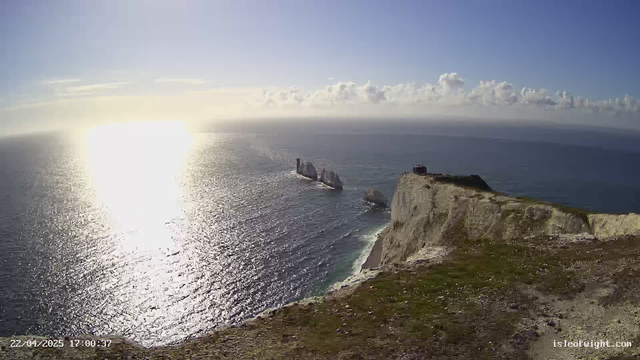 A scenic view of the ocean captured from a high cliff. The sun is shining brightly, reflecting off the water and creating a shimmering effect. There are a few small rock formations visible in the water, and a distant lighthouse can be seen near the larger rocks. The sky is mostly clear with some clouds, and the landscape features a grassy area at the cliff's edge leading down to the shore. The date and time are displayed in the lower left corner.