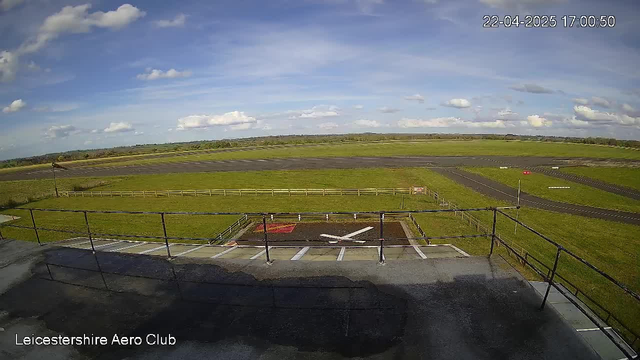 A wide, open landscape featuring a grassy airfield under a blue sky with scattered white clouds. In the foreground, there is a flat rooftop railing. Below, a helipad marked with a large white cross is visible, surrounded by a red area. In the distance, a runway stretches across the field, bordered by a wooden fence, with a patch of green grass on either side. The scene is well-lit and clear, indicating a sunny day.
