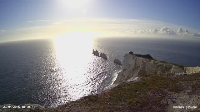 A scenic view of the ocean with sunlight reflecting off the water, creating a shimmering path. In the foreground, grassy cliffs extend to the edge where the land meets the sea. To the right, dark rock formations jut out of the water. A building is positioned near the cliff's edge, with a clear blue sky and scattered clouds above. The overall atmosphere conveys tranquility and natural beauty.