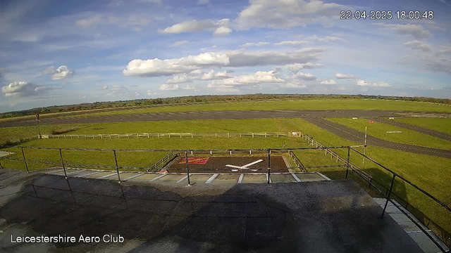 A view from a webcam overlooking Leicestershire Aero Club. The foreground features a fenced area with a white cross marking a landing or take-off spot on the ground. Beyond the fenced area is a wide expanse of green grass, with a few trees in the distance. The sky is mostly clear with a few fluffy white clouds, and the time displayed is 18:00 on April 22, 2025. The runway extends into the background, with several painted markings on it.