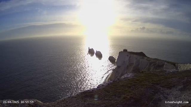 A panoramic view of the sea at sunset, with bright sunlight reflecting on the water surface, creating a shimmering effect. In the foreground, rugged cliffs descend towards the ocean, and three large rock formations are visible in the water. The sky is partly cloudy, with a soft gradient transitioning from blue to a warm orange hue near the horizon. The date and time are displayed in the bottom left corner of the image.