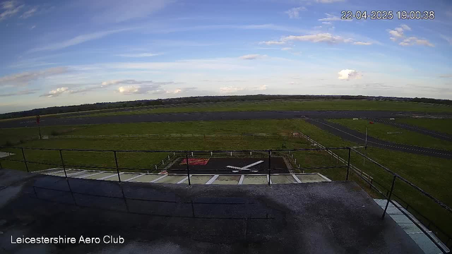 A clear view from a high balcony overlooking a grassy airfield. In the foreground, there is a black landing area marked with a white airplane symbol. The airfield is bordered by a low wooden fence, and the runway extends into the distance. The sky is mostly clear with a few scattered clouds, and the scene is bathed in soft evening light. The date and time are displayed in the corner.