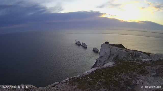 A picturesque view of the coastline featuring large white chalk cliffs on the right, with green grass and rocky terrain at the top. In the distance, several rocky formations protrude from the sea, some with a light structure on one. The sky is partly cloudy, with hues of blue and orange as the sun sets on the horizon, casting a gentle reflection on the calm water.