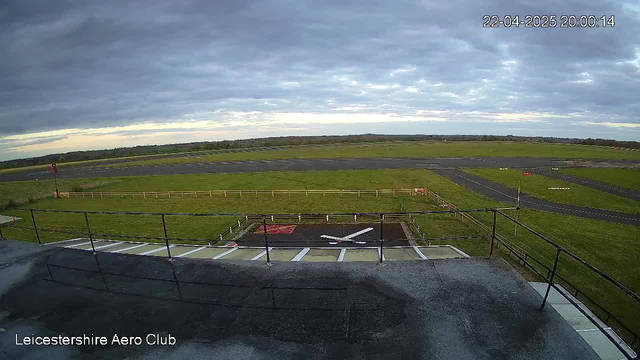 A webcam view of an airport environment under overcast skies during the evening. In the foreground, there is a railing with a gray surface below. The view shows a wide expanse of grass and a runway that leads into the distance, lined with a wooden fence on one side. To the left, a red light is visible, and there are runway markings on the tarmac, including a large white cross on the ground. The sky is filled with clouds, creating a muted atmosphere. The timestamp in the corner indicates the date and time.