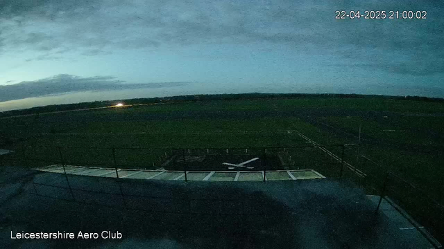 A dimly lit landscape at dusk shows a wide, open field and a runway at the Leicestershire Aero Club. The sky is filled with gray clouds, and a faint light can be seen in the distance, indicating perhaps an aircraft light or headlights. In the foreground, the edge of a building with a flat roof is visible, along with a white airplane silhouette on the ground near the runway. The overall scene conveys a serene atmosphere as day transitions to night.