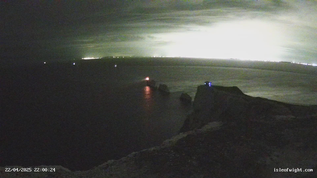 A night view of a coastline with dramatic cliffs and a dark ocean. In the foreground, rugged rocks are visible, while a lighthouse emits a red light on one of the nearby rocks. Multiple small boats with lights can be seen in the water. The background features a distant glow from the town or city lights shimmering against a cloudy sky, creating a contrast against the darkness of the sea.