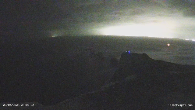 A dimly lit coastal scene is visible, with a prominently silhouetted rocky outcrop on the right. In the background, a faint glow of lights from a distant shoreline can be seen, suggesting the presence of a town or city. The sky is overcast, and the overall atmosphere is dark and moody. There are subtle hints of waves or water in the foreground, blending into the dark surroundings. A blue light is noticeable on the outcrop, adding a hint of color to the otherwise muted image. The date and time are displayed in the bottom left corner.