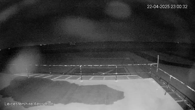 A dark scene captured from a webcam. The foreground shows a flat surface with some markings, possibly indicating a runway or parking area for aircraft. A metal railing runs along the edge of the area, and in the distance, faint lights can be seen on the horizon, suggesting the presence of buildings or vehicles. The overall atmosphere is dimly lit, reflecting a nighttime setting. The timestamp in the corner indicates the image was taken on April 22, 2025, at 11:00 PM.