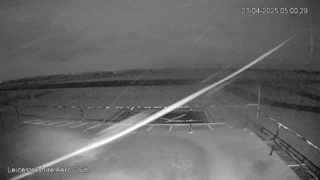 A dimly lit image taken from a webcam showing a flat landscape with a few outlined features in the foreground. In the foreground, there is a railing or fence, and a section of a runway or parking area is visible. The background features a cloudy sky, indicating low visibility, and the scene appears monochromatic with shades of gray. The time and date stamp at the top right reads April 23, 2025, at 5:00 AM.