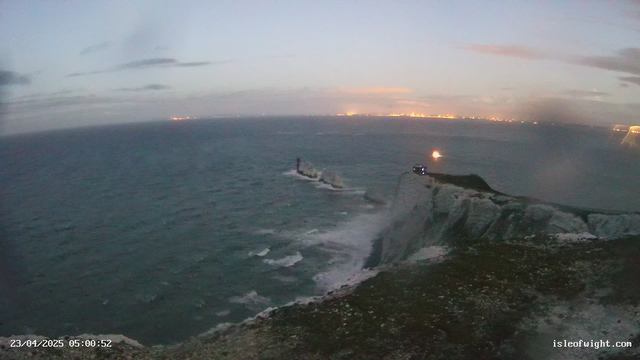 A coastal scene at dawn featuring rough waves crashing against a cliff. A lighthouse is partially visible to the right on a rock formation, with a light beacon shining. The sky is gray with scattered clouds, and the distant horizon shows twinkling lights from a town. The foreground includes rocky terrain and a small structure near the cliff's edge.