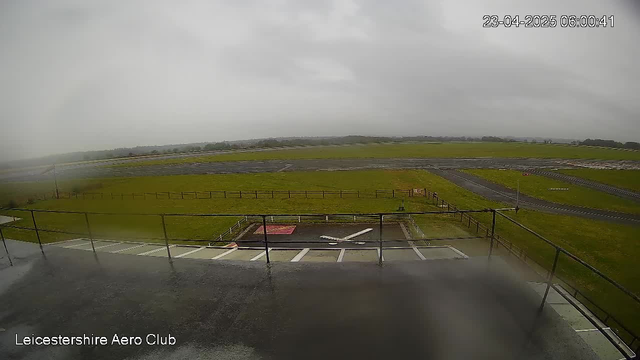 A view from a webcam at Leicestershire Aero Club showing a wet runway under overcast skies. The foreground features a railing and pavement with a white cross marking an area, while the background displays a grassy field and a portion of the runway. The scene appears gloomy, with fading visibility.