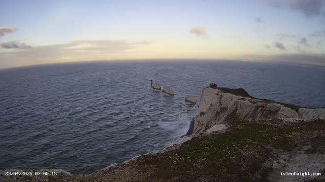 A panoramic view of the ocean at dawn, with gentle waves lapping against a rocky shoreline. In the foreground, there is a cliff with green grass and scattered rocks. In the midground, several weathered rock formations protrude from the water. A red structure is visible on one of the rocks, and another structure is perched on top of the cliff. The sky is filled with soft, pastel clouds and light blue hues, suggesting early morning light.