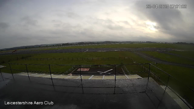 A cloudy morning view from the Leicestershire Aero Club webcam, showing a wide expanse of grass and a runway. There is a small white aircraft on the ground with a runway marking nearby. In the background, a fence lines the area with additional runway surfaces visible. The sky is overcast with vague hints of brighter clouds near the horizon. The timestamp in the corner indicates the image was captured on April 23, 2025, at 7:01 AM.