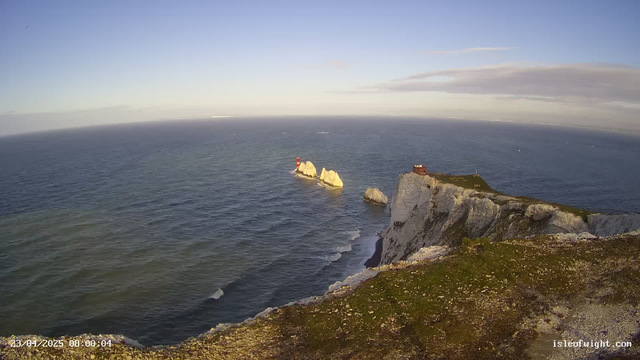 A coastal landscape featuring a rocky cliff that descends into the sea. In the foreground, there is vegetation on the cliff's edge, leading to steep slopes. Below, waves gently crash against the rocks. In the distance, two prominent white chalk formations rise from the water, with a red lighthouse on one of them. The sky is clear with some scattered clouds, and a light blue hue dominates the scene. The horizon stretches far into the ocean, indicating a calm day.