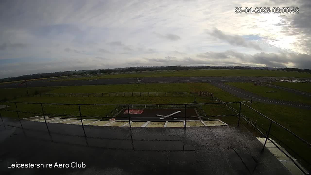 A view from a webcam at Leicestershire Aero Club shows an outdoor scene with a runway and green fields. The sky is overcast with scattered clouds, and a wet surface suggests recent rain. In the foreground, there is a small area with a red square and a white object resembling an aircraft. The horizon stretches into the distance with fields and signs of fencing. The date and time displayed in the corner indicate it is early morning.
