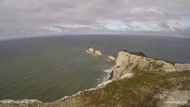 A coastal view featuring white chalk cliffs rising sharply from the sea. In the foreground, there's greenery and rocky terrain. To the right, a small red lighthouse stands on a rocky outcrop, accompanied by another rock formation. The ocean extends into the distance, with gentle waves and a cloudy sky above, indicating a breezy day.