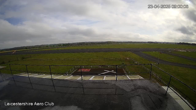 A view from the Leicestershire Aero Club webcam shows a wide open field under a cloudy sky. In the foreground, a flat rooftop with a railing is visible. Below, there is an area of grass encompassing a large landing area marked with a white cross, and a section displaying a red surface. The landscape is mostly green, with an outlined runway in the distance, bordered by some fencing and scattered trees. The time stamp in the corner indicates the date and time of the image.