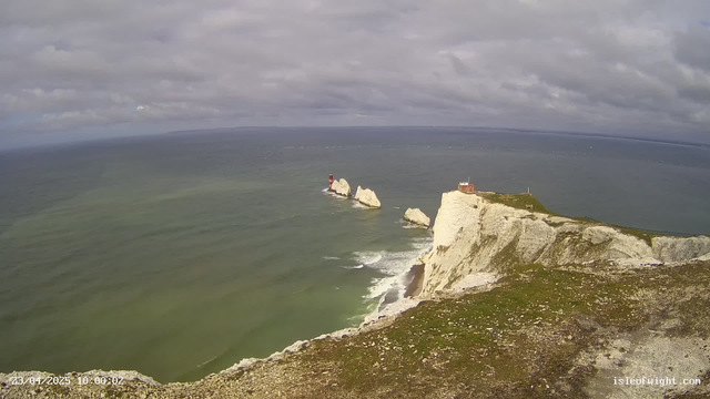 A coastal scene featuring rugged white cliffs and a calm greenish-blue sea under a partly cloudy sky. In the distance, two rocky outcrops emerge from the water, with a red lighthouse positioned on one of them. The foreground shows rocky terrain on the cliff edge with sparse grass.