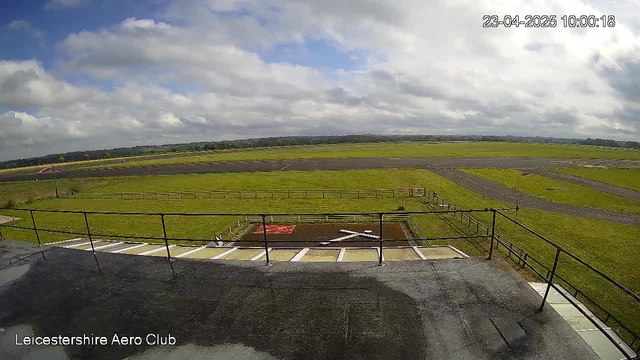 A view from the top of a building at the Leicestershire Aero Club, looking out over a grassy airfield. The sky is mostly cloudy with patches of blue. In the foreground, there is a railing around the edge of the building. Below, a grassy area is marked with a red and white cross. The airfield stretches out with a paved runway visible in the background surrounded by more grass and a few scattered fences.