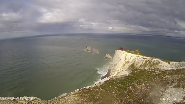 A coastal view featuring white chalk cliffs dropping into the blue sea. In the foreground, a grassy area with some rocky textures. Two white rock formations are visible in the water. A small red building is located at the top of the cliffs, while another red structure appears on one of the rock formations. The sky is mostly cloudy with varying shades of gray and blue above the horizon.
