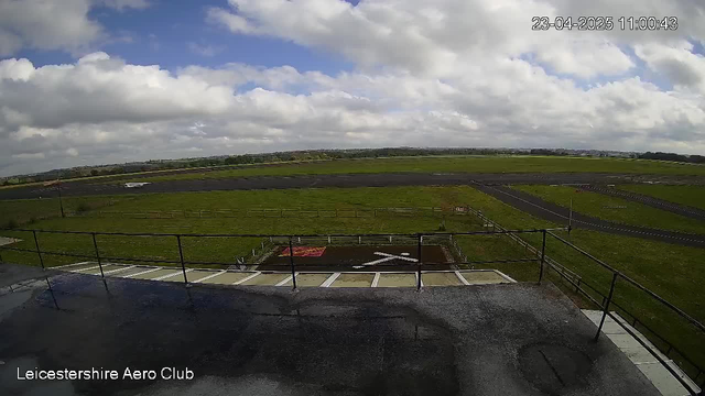 A view from the Leicestershire Aero Club webcam shows a wide, open grassy area with a tarmac runway. The sky is mostly cloudy, with patches of blue peeking through. In the distance, there are rolling green hills. On the runway, a small aircraft is visible, positioned away from the camera. In the foreground, a flat surface with light reflections and railings can be seen, framing the view of the runway and landscape.