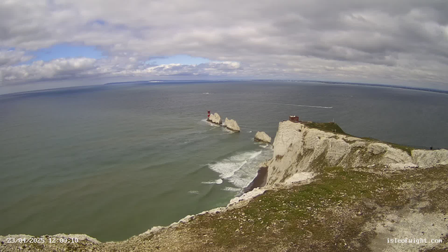A scenic view of a coastal landscape featuring white chalk cliffs, lush green grass, and the calm blue sea. In the distance, several rocky formations emerge from the water, with a red lighthouse on one of the rocks. The sky is partly cloudy with patches of blue visible. The image captures a tranquil shoreline where gentle waves lap against the cliffs.