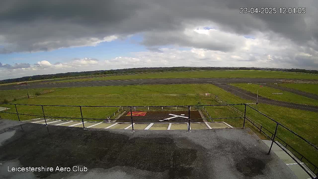 A view from a raised platform at the Leicestershire Aero Club, showing an expansive green field with a runway in the background. The sky is partly cloudy with patches of blue peeking through. A visible white cross marking is located on the ground, and there are fences along the edges of the field. The image captures a serene outdoor scene with no aircraft present.