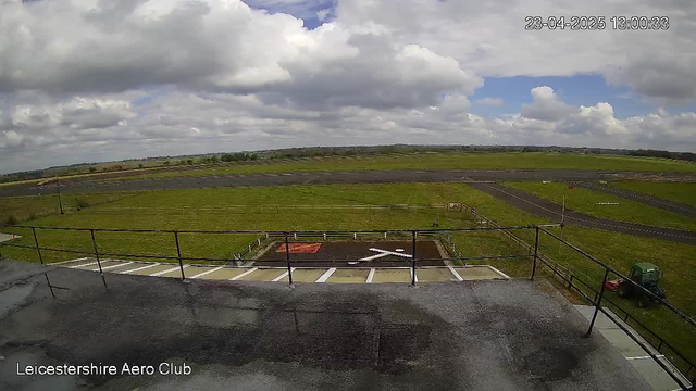 A wide view of an airport runway with grassy fields surrounding it. In the foreground, there is a helipad marked with a white 'X' on a red background. The sky is partly cloudy with blue patches visible. A tractor can be seen in the distance on the right side of the image. The airport area appears well-maintained with a few trees and fences in the background.