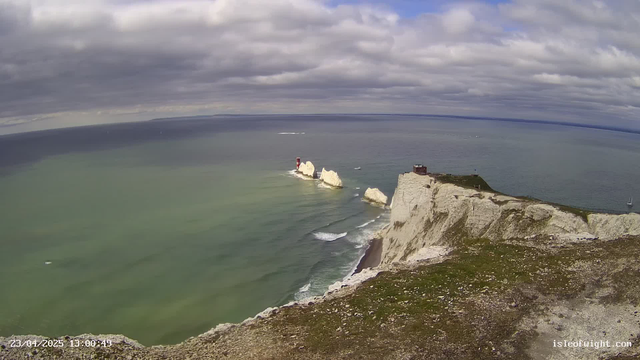 A coastal view featuring white chalk cliffs with some grassy areas at the top. In the water, there are a few small boats, and further out at sea, there are additional sailing vessels. The sky is predominantly cloudy with hints of blue peeking through. At the base of the cliffs, there are waves gently lapping against the shore. In the distance, distinctive red and white lighthouses are visible on the cliff formations.