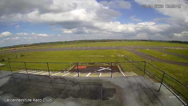 A wide view of an airfield is presented, showing a grassy area and a runway in the distance. The sky is partly cloudy with patches of blue. In the foreground, there is a marked area with a red flag and an "X" shape on the ground. A railing is visible along the bottom edge of the image, and a sign can be spotted to the right. The scene conveys a tranquil outdoor setting at the Leicestershire Aero Club.