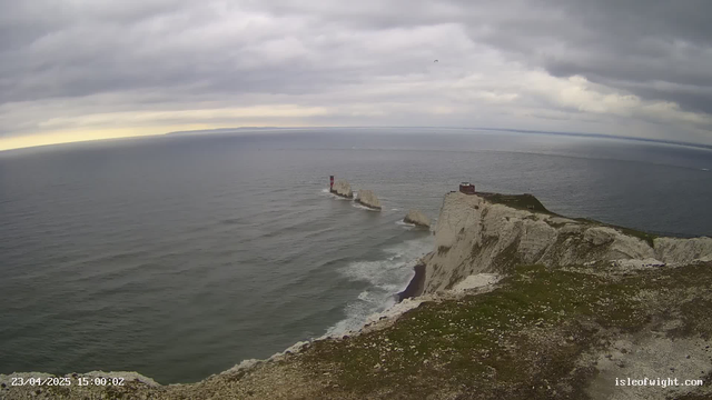 A coastal scene shows a rocky shoreline under a cloudy sky. The ocean is calm with gentle waves. In the center, two rocky outcrops rise from the water, topped with white cliffs. A red lighthouse stands on one of the outcrops. In the foreground, green grass covers the cliff's edge, and the rugged terrain is visible. The horizon shows lighter clouds, hinting at the sun behind them.