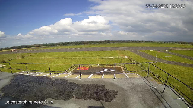 A view from a webcam at Leicestershire Aero Club, showing a spacious grassy area under a bright blue sky with fluffy white clouds. In the foreground, there is a marked landing spot with a white cross on a dark surface, and nearby, a fenced area with a flag pole and a distant runway. The landscape is predominantly green with sections of grey asphalt and a lightly rolling horizon.