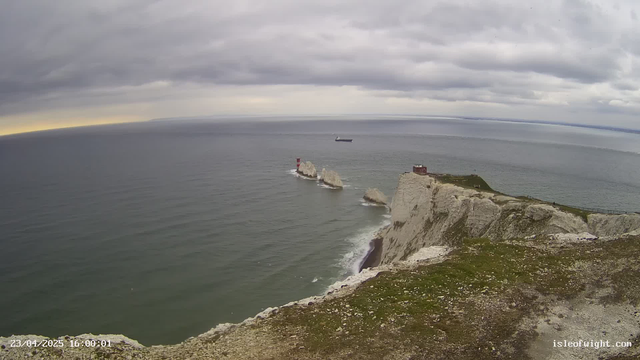 A panoramic view of a coastal scene featuring cliffs with a grassy top. The ocean is calm, with a few small waves. A large cargo ship is visible in the distance, sailing across the water. To the left of the image, there are three rock formations with a red and white striped lighthouse on one of them. The sky is overcast with gray clouds, and there is a hint of brighter light on the horizon.