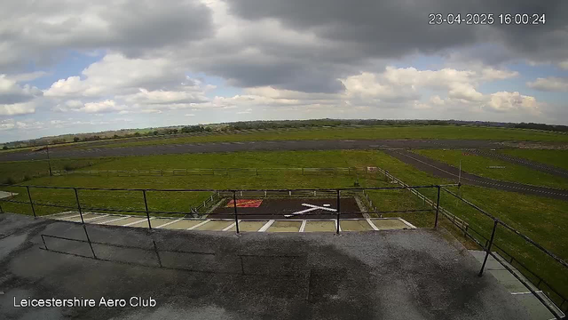 A view from a webcam overlooking an airport setting. In the foreground, a concrete terrace is visible with a railing. Below the terrace is a grassy area with a white cross marking on the ground. The background features a long runway with a few parallel lines stretching across the green landscape. The sky is partly cloudy with patches of blue visible among the clouds, indicating a bright but overcast day. The date and time are displayed in the corner.