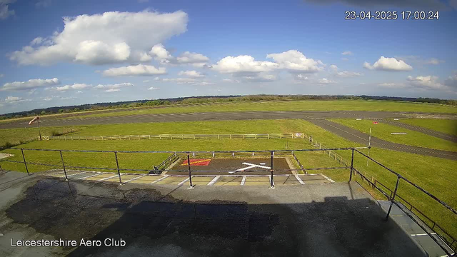 A view from a webcam at Leicestershire Aero Club, capturing a bright and clear sky with fluffy white clouds. Below, an expansive green field stretches out, intersected by a grey runway. In the foreground, there's a red area marked with a white X, and a railing along the edge of a building. A flag is visible on the left side, gently fluttering in the wind. The farthest background includes more open fields and trees against a distant blue sky.
