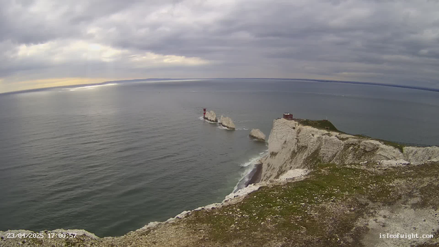A coastal view showing white chalk cliffs descending into the sea, with calm water extending to the horizon. A cloudy sky above with some breaks of light. In the distance, there are three distinct rock formations, two of which have red and white striped lighthouses on them. A small building is positioned at the edge of the cliff on the right side of the image. The foreground features grass and rocky terrain.