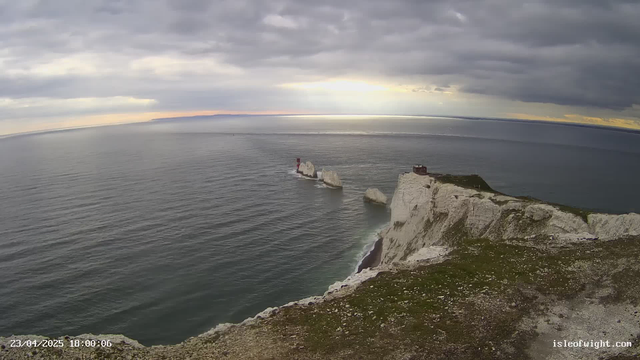 A coastal scene featuring steep white cliffs on the right side, leading down to the ocean. In the distance, a lighthouse with a red and white striped design stands on a rocky outcrop in the water. The sky is overcast with varying shades of gray, and beams of sunlight break through the clouds near the horizon, illuminating the water's surface. The foreground includes grassy land with scattered rocks. The ocean appears calm with gentle waves.