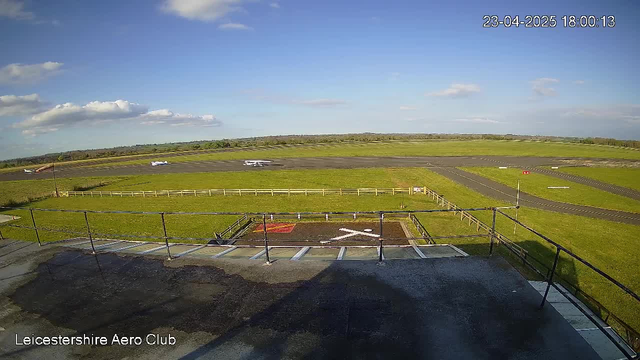 A clear view of an airport runway and surrounding landscape during the day. The foreground features a rooftop with railings, while a red and white marked area is visible on the ground below. In the background, there are grassy fields, a clear sky with some clouds, and several small aircraft parked on the runway. The overall scene is bright and well-lit, indicating a sunny day.