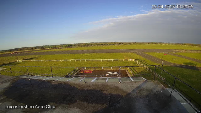 A view from a webcam overlooking Leicestershire Aero Club. The image shows a large, open space with a runway stretching across the center, bordered by green grass. In the foreground, there is a fenced area with a red patch and a white cross marking a designated spot. A light breeze movement is indicated by a flag situated to the left. The sky is mostly clear with a few clouds, and the image captures a tranquil atmosphere at sunset.