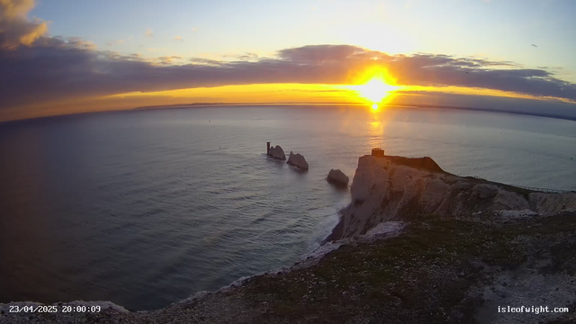 A picturesque sunset over the ocean, with vibrant orange and yellow hues illuminating the sky. The sun is low on the horizon, casting a warm glow on the water's surface. In the foreground, there are rugged white cliffs and several large rocks protruding from the sea. A lighthouse-like structure is visible atop one of the cliffs, and a path can be seen winding along the coastline. The scene conveys a sense of tranquility and natural beauty.